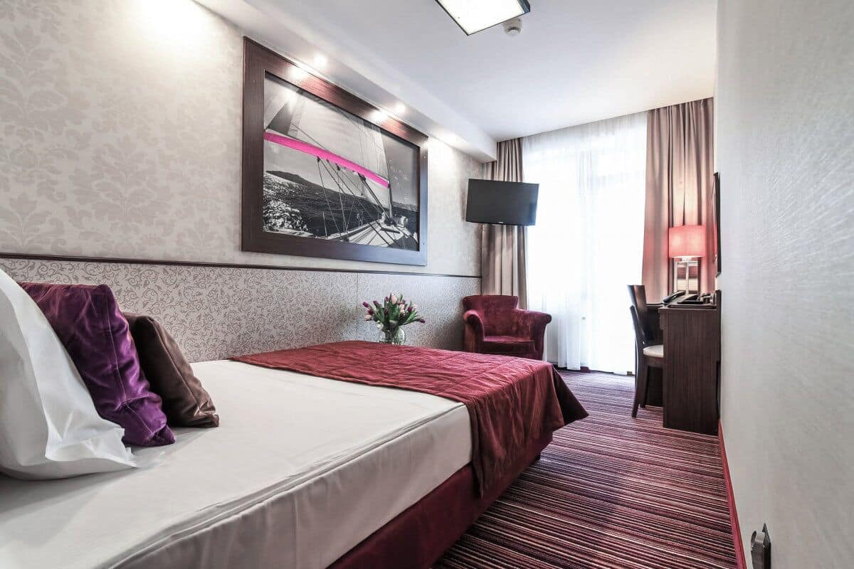 A hotel room with a bed, a desk, a chair, a TV, and a large window with curtains.