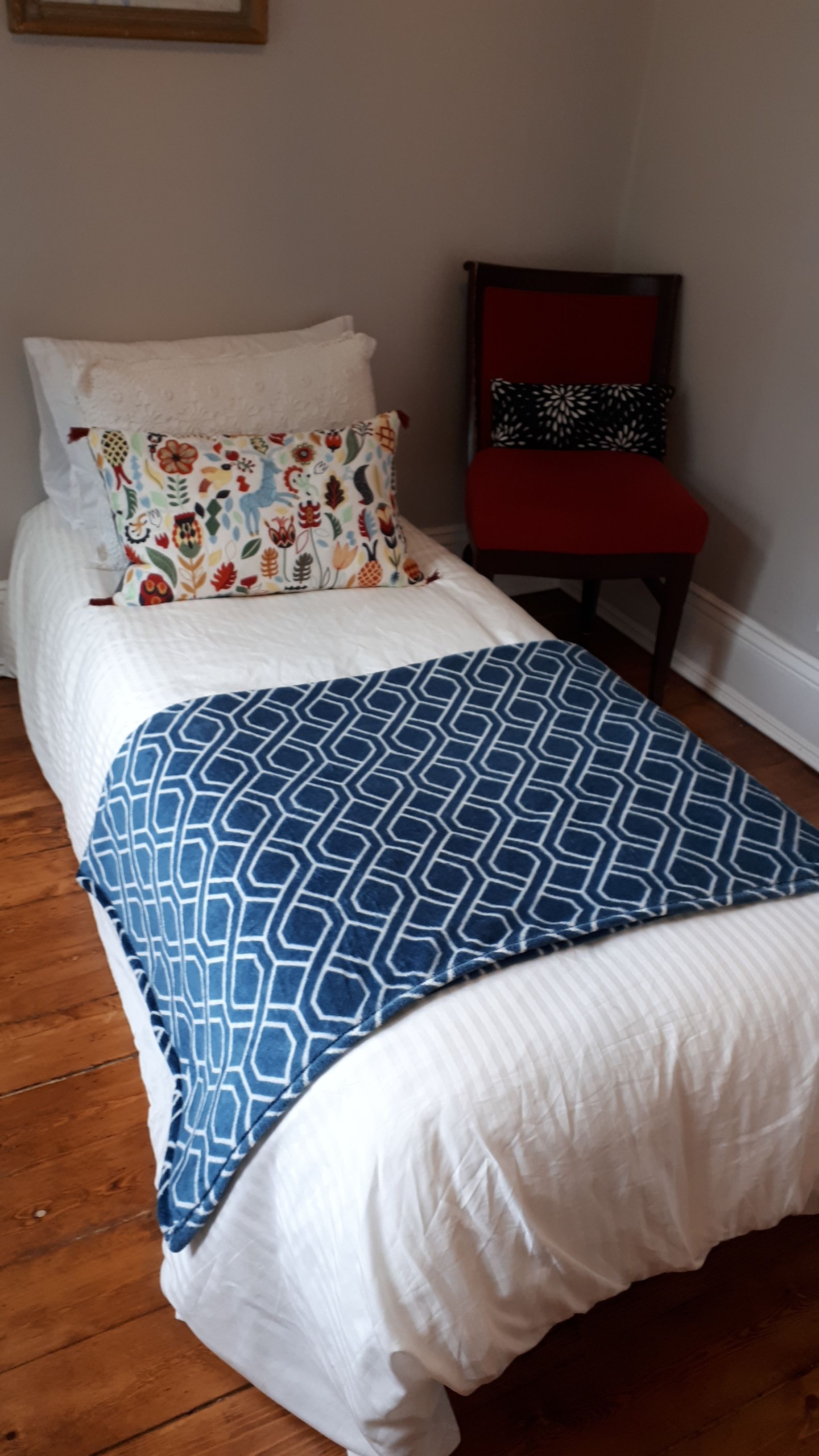 Un letto con copriletto bianco e blu decorato con motivi, un cuscino floreale e una sedia rossa con cuscino nero a fantasia.