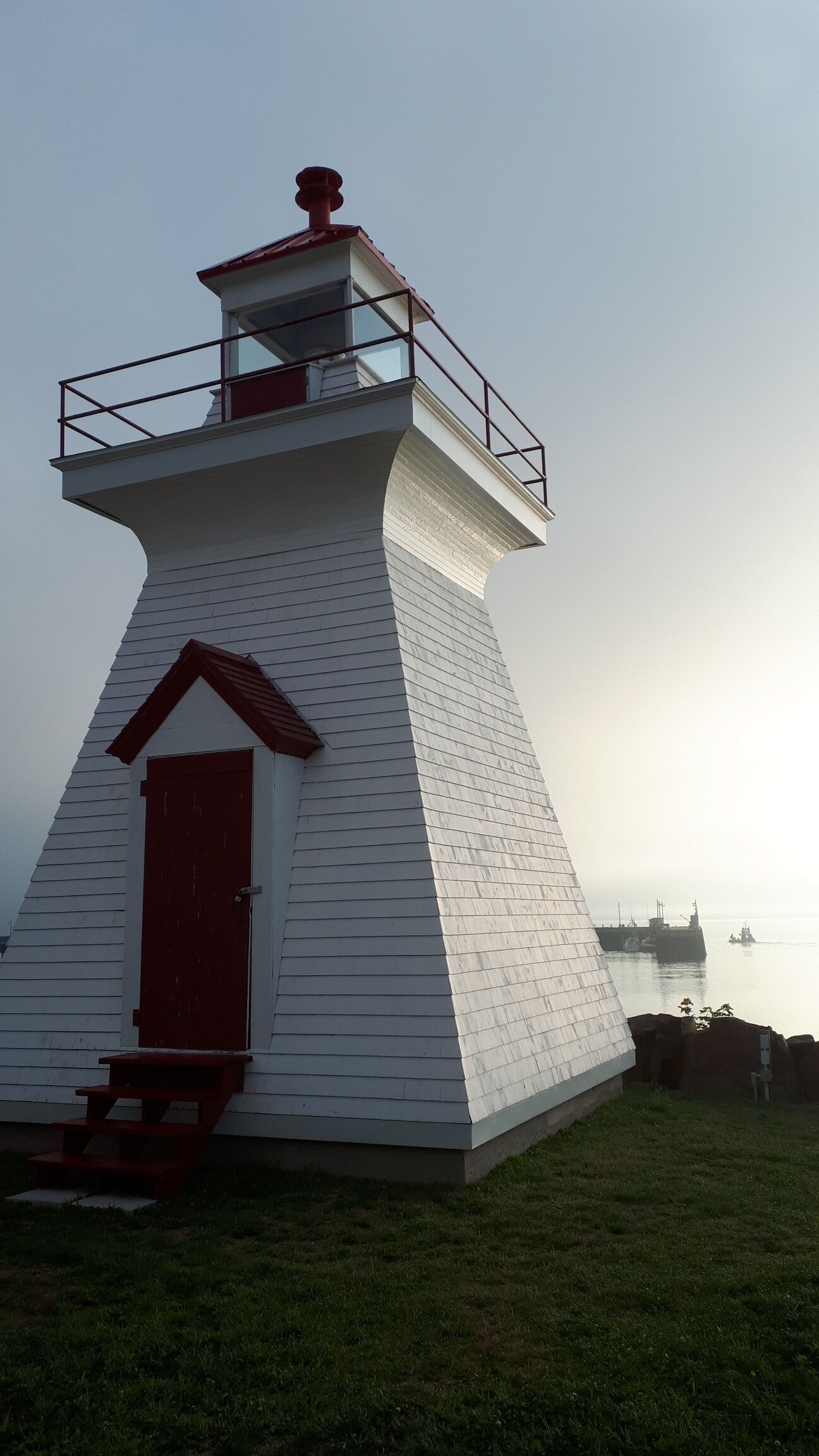 Un phare à l’extérieur blanc et à la porte rouge, situé sur une zone herbeuse près d’une étendue d’eau.
