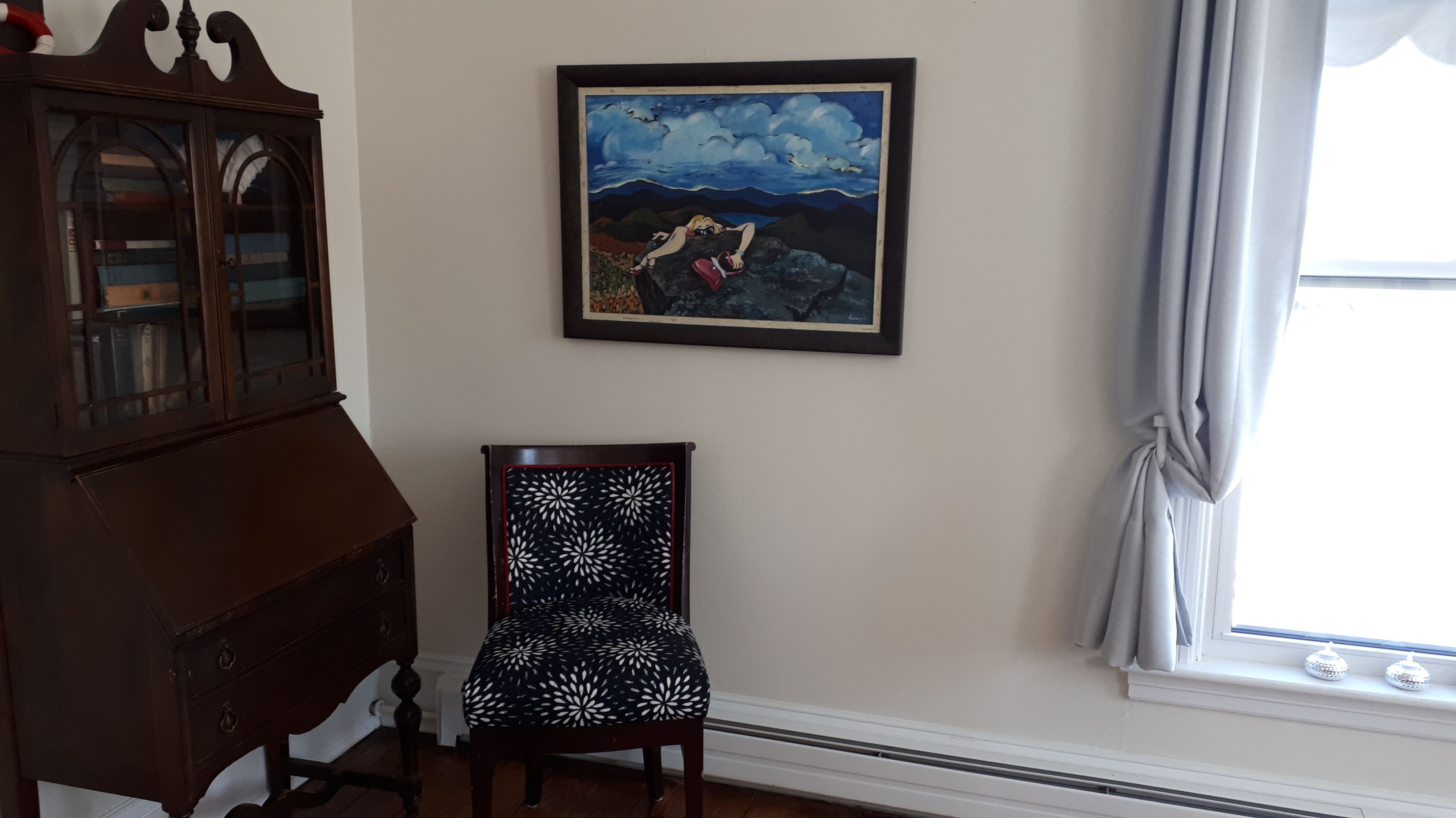 A room with a dark wooden cabinet, a chair with a black and white patterned cushion, and a framed painting of a landscape with a figure climbing a rock.