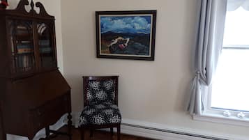 A room with a dark wooden cabinet, a chair with a black and white patterned cushion, and a framed painting of a landscape with a figure climbing a rock.