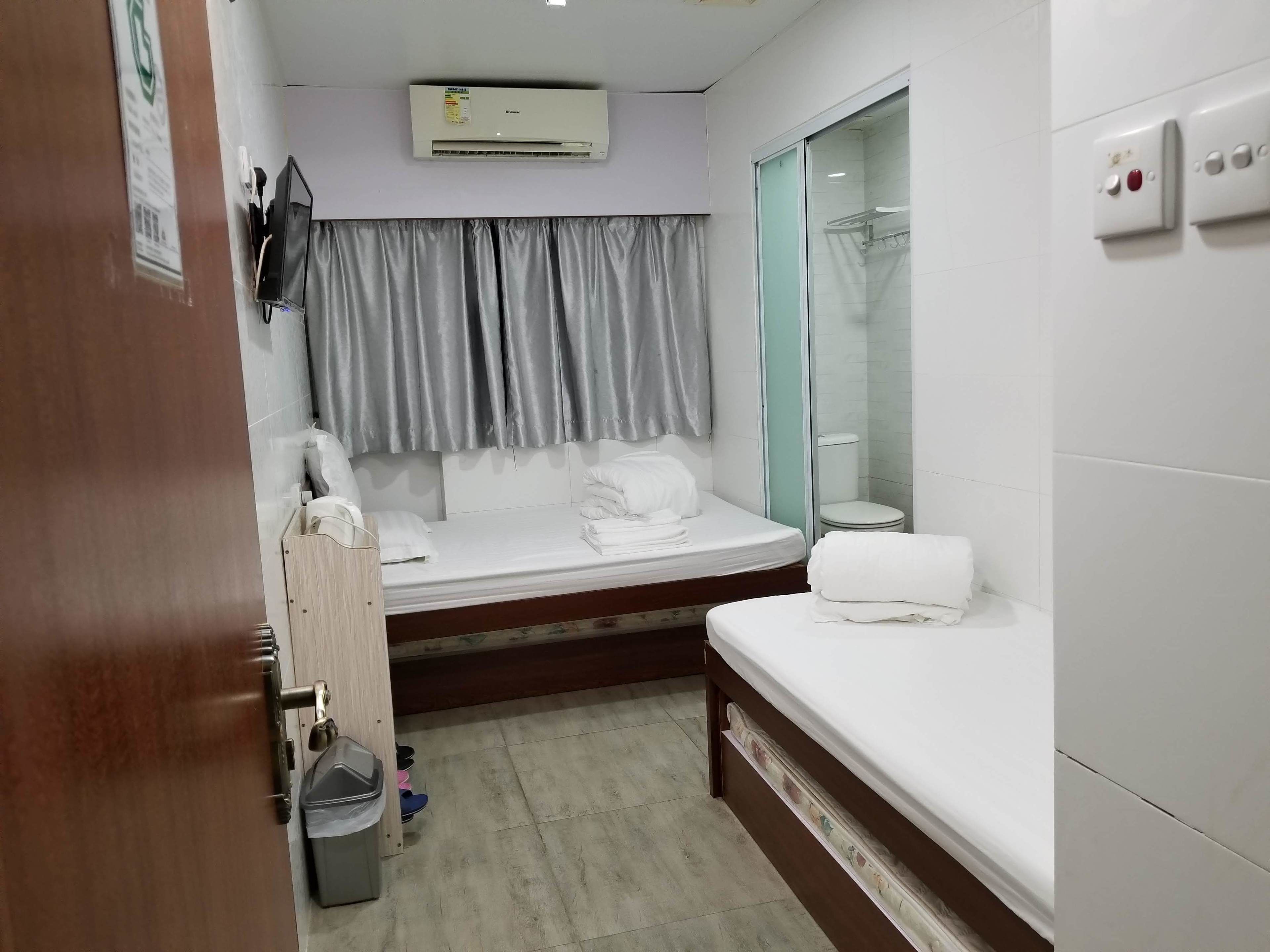 A compact hotel room with a bed, a small bedside table, a trash bin, a window with curtains, an air conditioning unit, and a shower area.