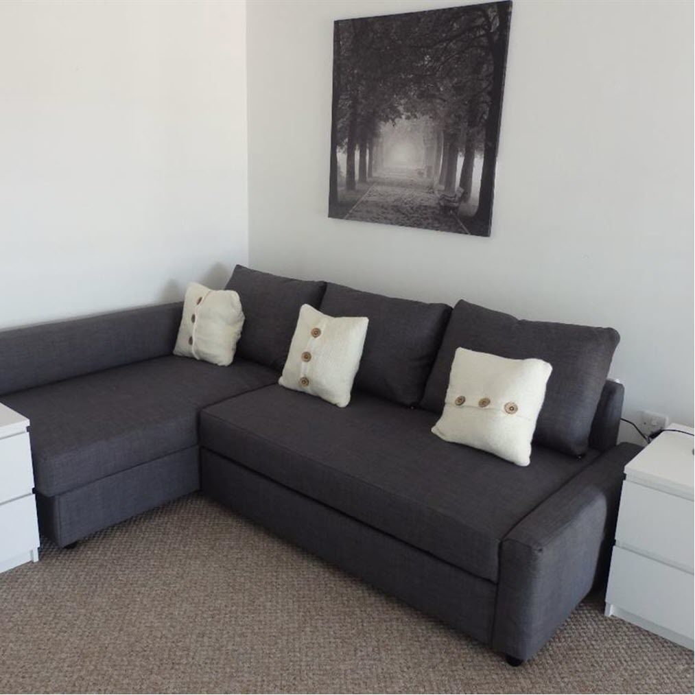 A grey sectional sofa with three cushions, a white side table, and a framed picture of a tree-lined path on the wall.