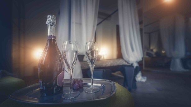 Una botella de champán, dos copas y uvas en una bandeja en una habitación con poca luz, con cama y cortinas.