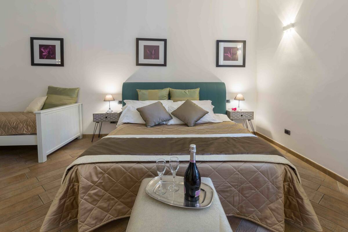 A hotel room with a bed, bedside tables, a bedside lamp, a tray with a bottle and glasses, and framed pictures on the wall.