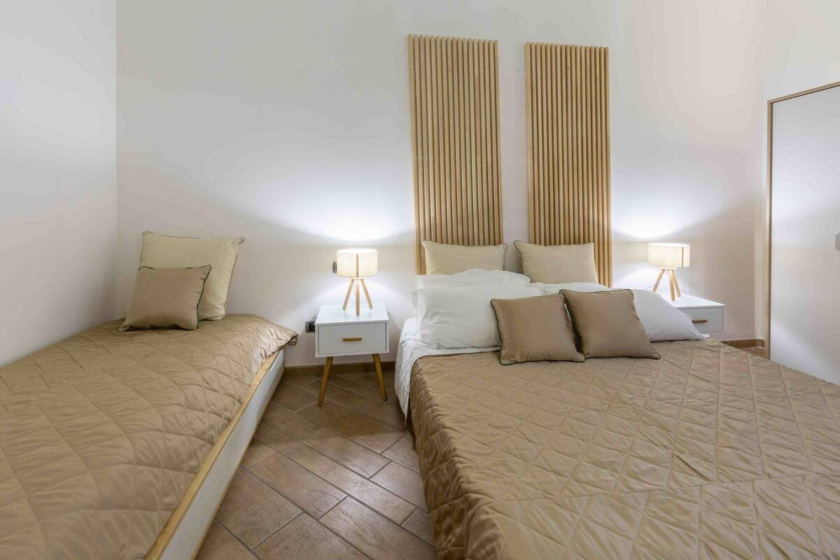 A hotel room with two beds, each with a beige quilted bedspread and matching pillows. There are bedside tables with lamps and a wooden headboard.