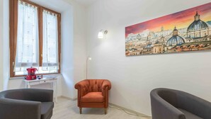 Una habitación con un ventanal grande, un cuadro de un paisaje urbano, dos sillones y una mesita con un hervidor rojo.