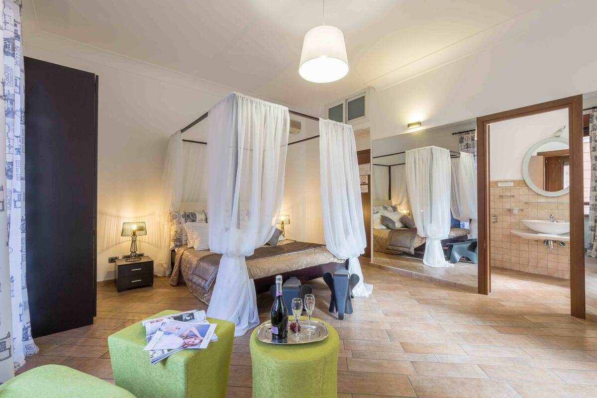 A bedroom with a four-poster bed, a bedside table with a lamp, and a green ottoman with magazines.