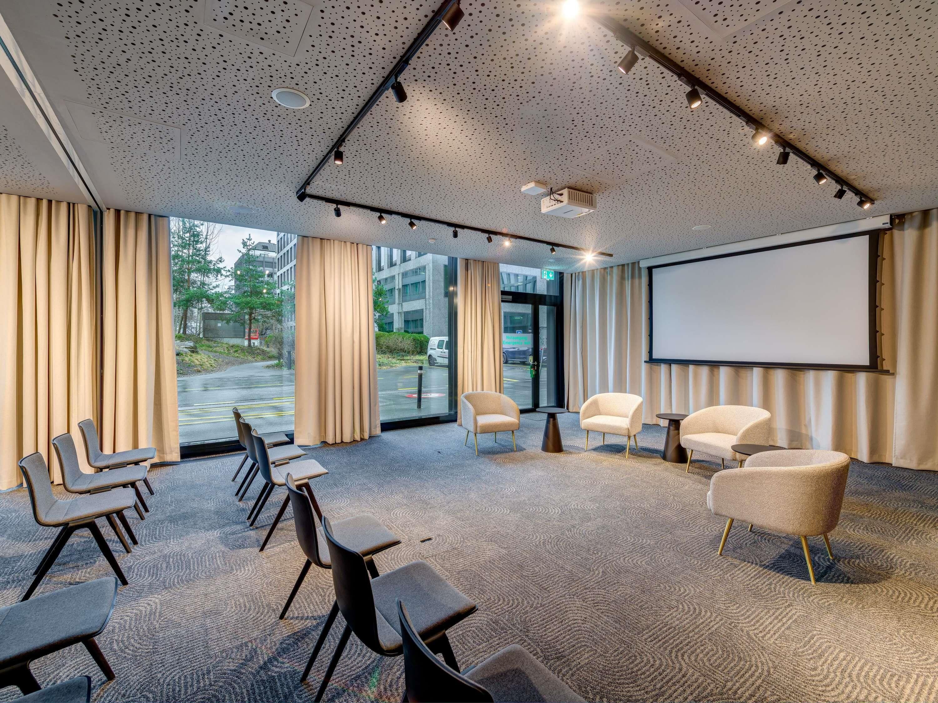 Un salón de conferencias con pantalla de proyector, un amplio ventanal con cortinas y suelo alfombrado.