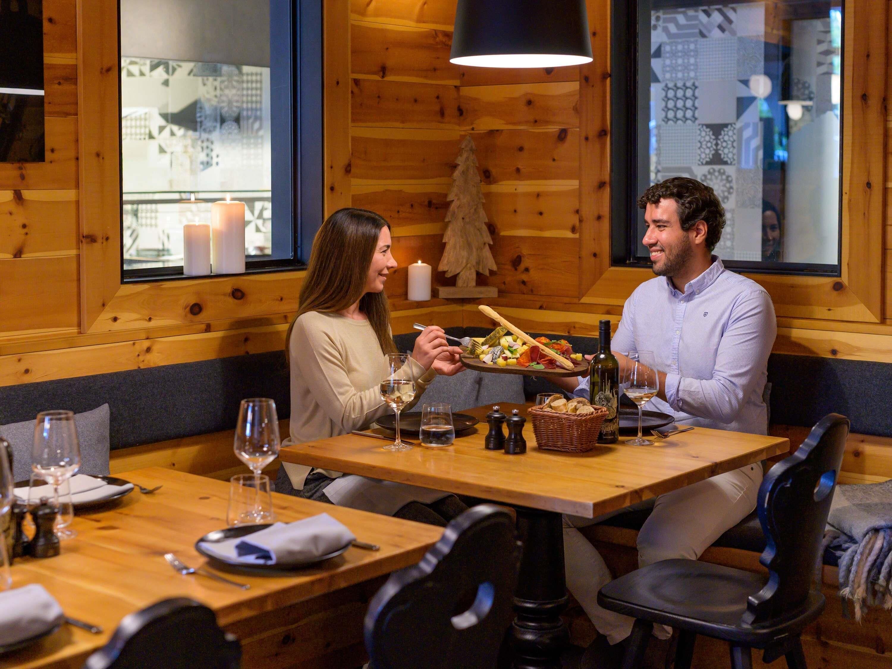 Een houten interieur met een eettafel gedekt voor twee personen, inclusief een fles wijn, een broodmand en een bord met diverse kazen en vleeswaren.