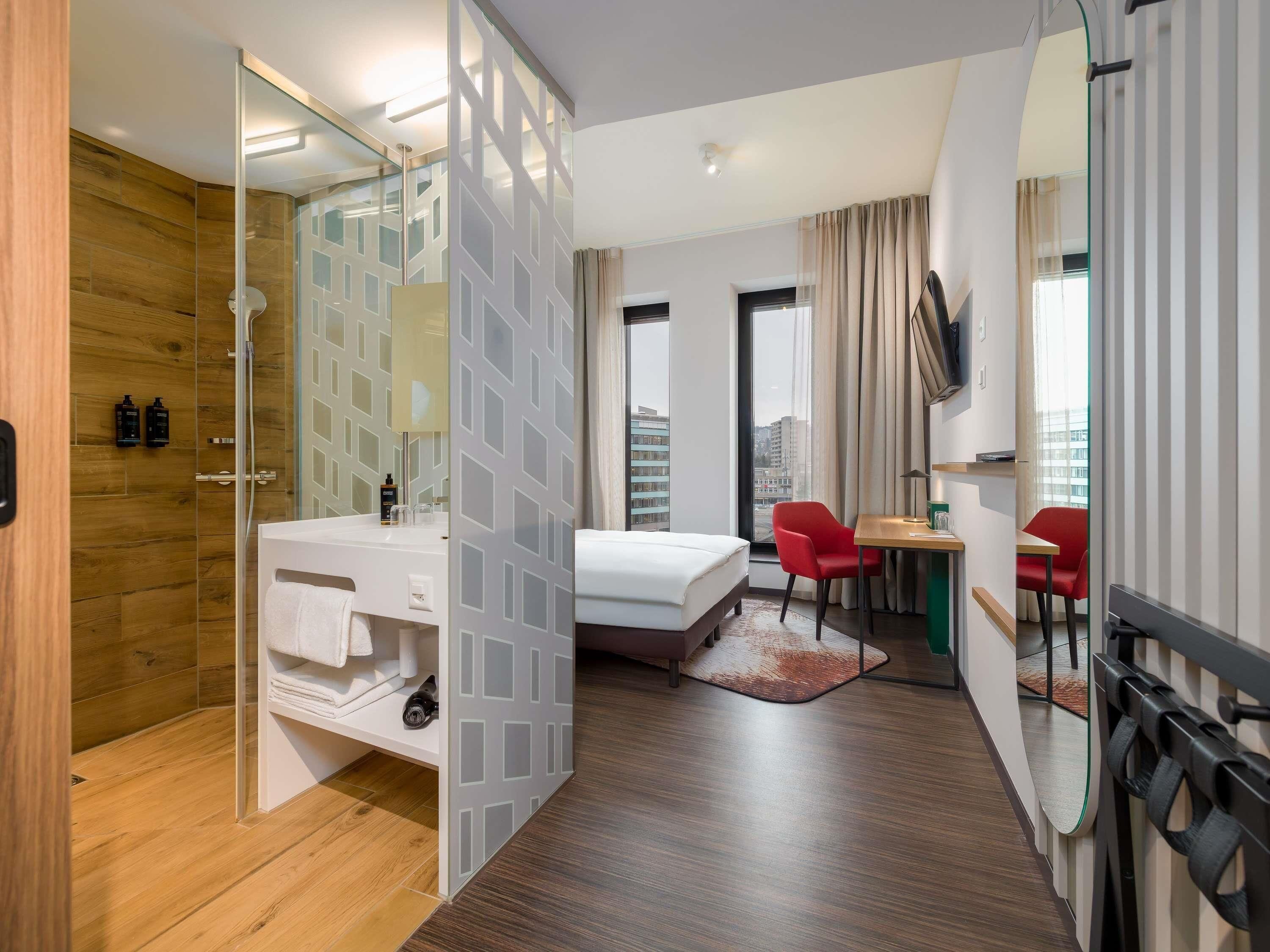 A hotel room with a wooden floor, a bed, a desk, a chair, a shower area, and a view of the city.