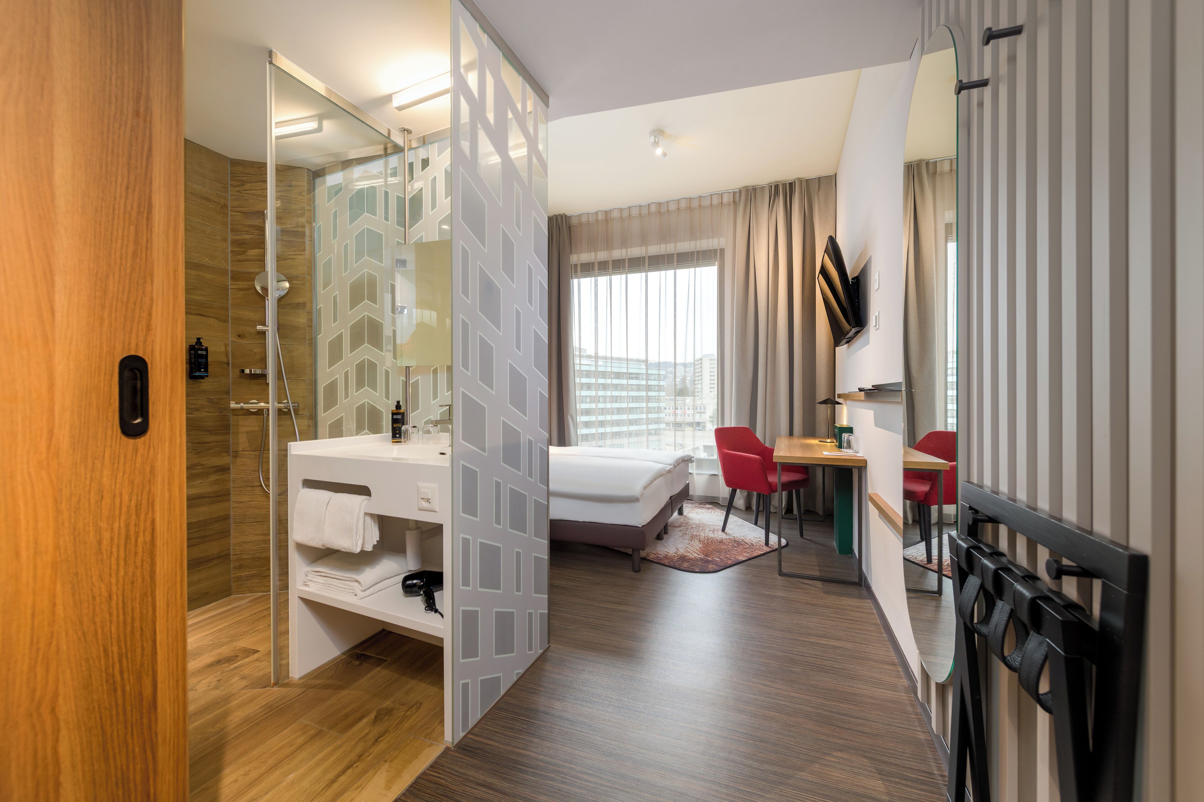 Habitación de hotel moderna con suelo de madera, cabina de ducha de vidrio, tocador blanco, cama, escritorio con silla y vista a la ciudad por la ventana.