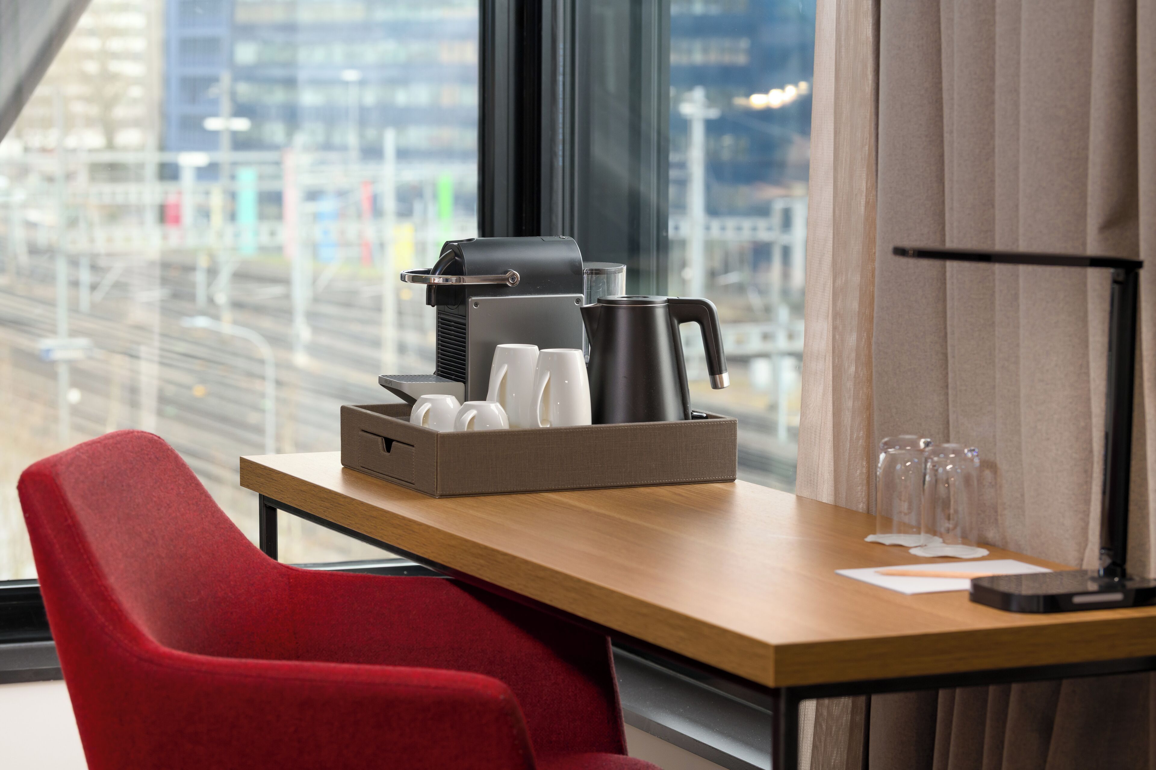 A hotel room with a wooden table, a red chair, a tray with coffee cups and a coffee pot, and a view of a cityscape through large windows.