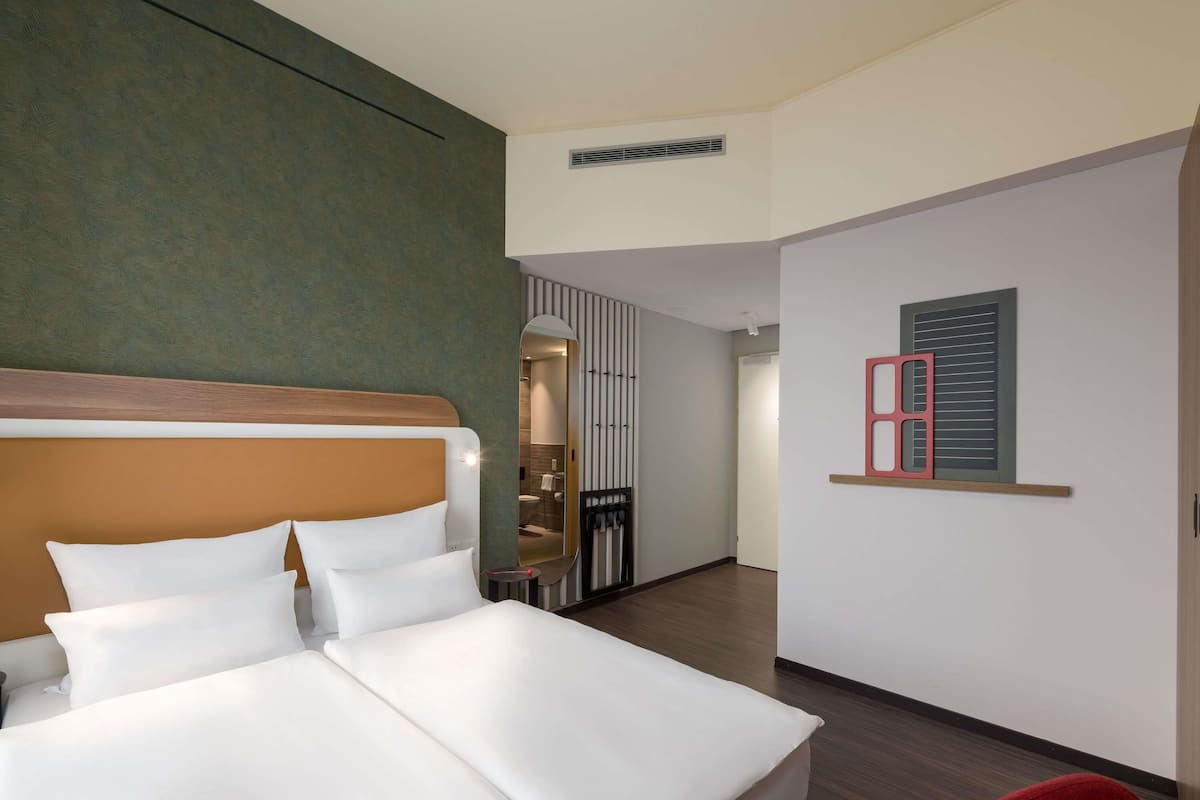Une chambre d’hôtel avec un lit, un miroir, une chaise rouge et une œuvre d’art accrochée au mur.