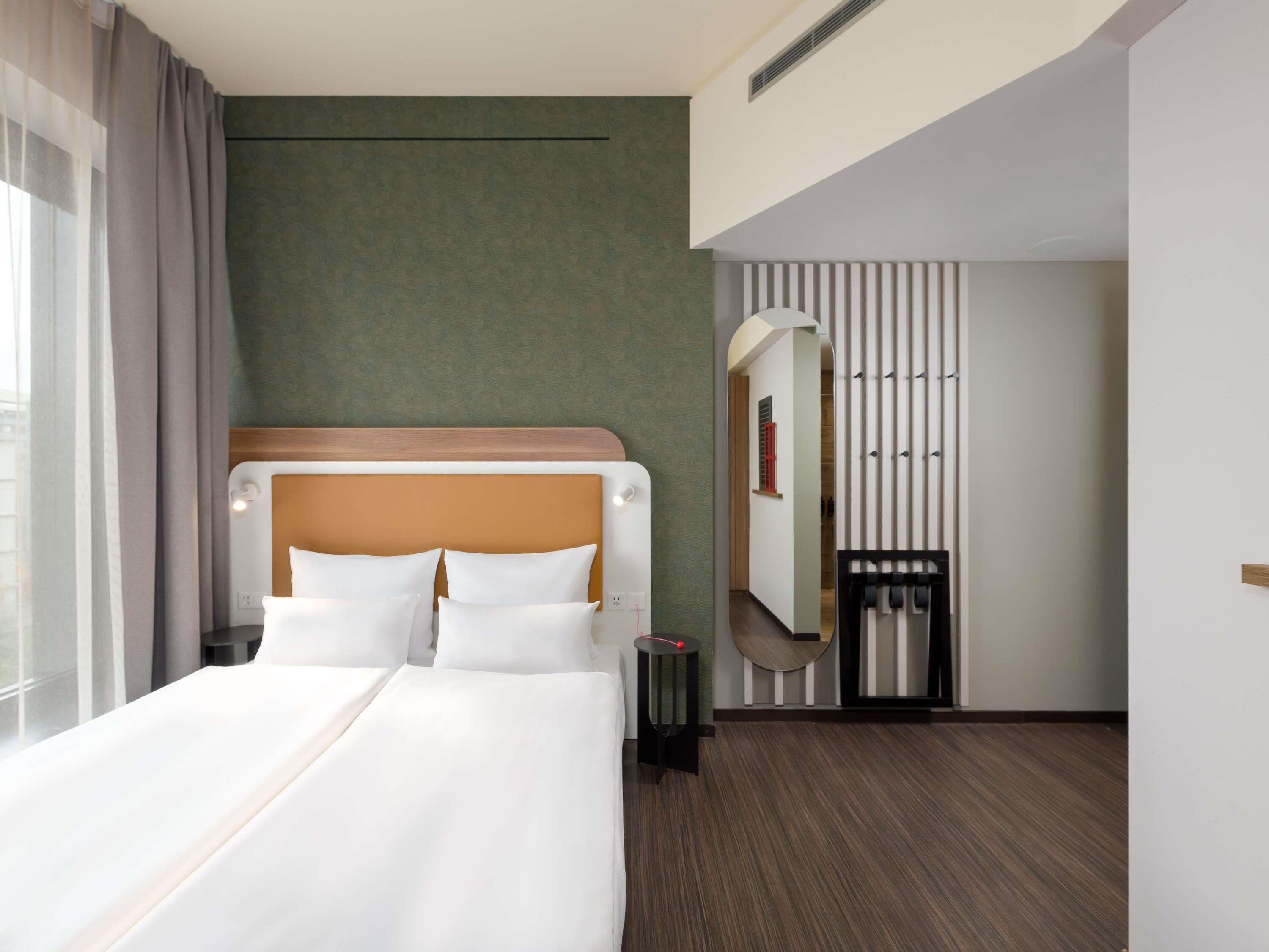 Una habitación de hotel con una cama grande, un cabecero de madera, una mesita pequeña, un espejo y una puerta con persianas verticales.