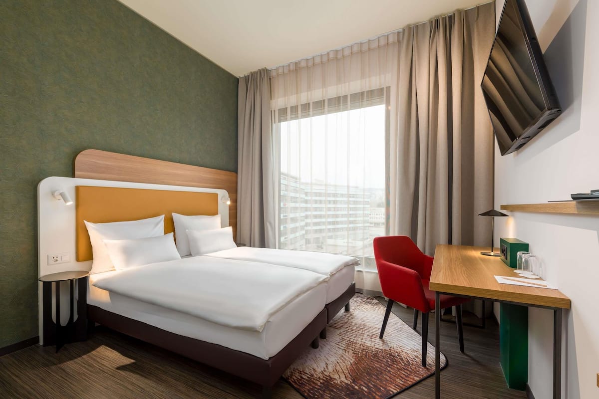 Une chambre d’hôtel équipée d’un lit, d’un bureau, d’une chaise, d’un téléviseur fixé au mur et d’une fenêtre avec des rideaux.