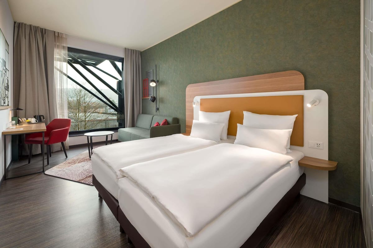 Une chambre d’hôtel avec un grand lit, un bureau avec une chaise, une petite table et une fenêtre avec des rideaux.