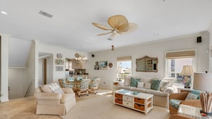 A spacious living area with a high ceiling fan, a dining table with chairs, a sofa set, and a large window with a view.