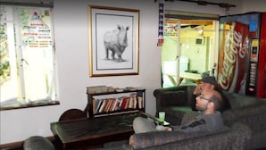 A living room with a sofa, a coffee table, and a bookshelf. There is a framed picture on the wall and a Coca-Cola vending machine. - Johannesburg Youth Hostel (Randburg)