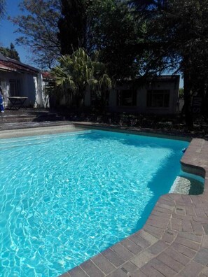 Una piscina azul cristalina con borde de ladrillo, rodeada de árboles y un edificio al fondo.