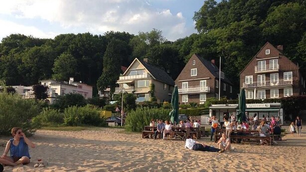 Una spiaggia con persone che si rilassano e cenano. Si vedono edifici sullo sfondo.