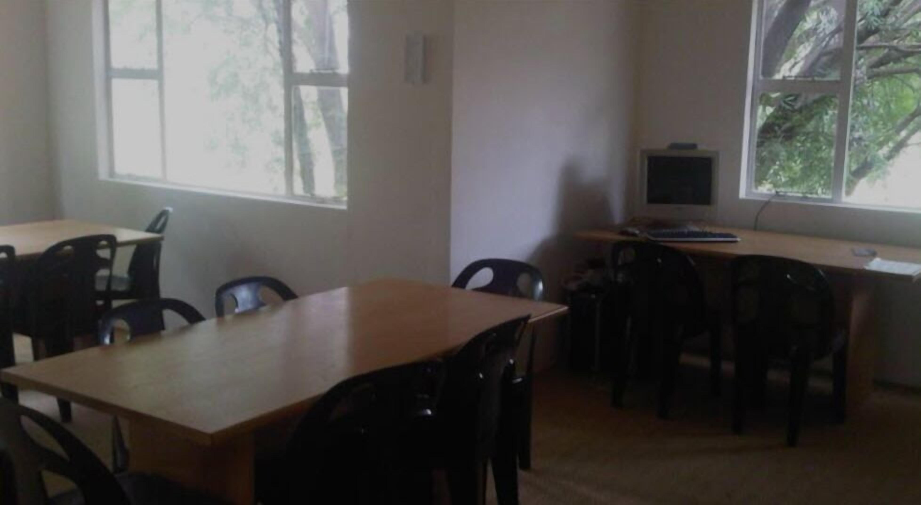 A classroom with wooden tables and black chairs, a television on a desk, and windows with a view of trees.