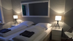 En dobbeltseng med hvide sengetøj, to sengeborde med lamper, en stol med hvid pude og et vindue med udsigt til træer.