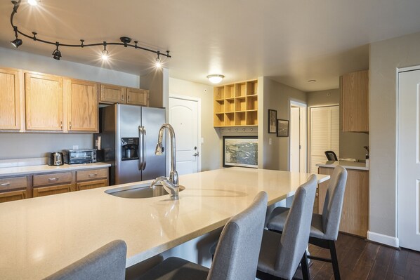 A modern kitchen with wooden cabinets, a stainless steel refrigerator, a built-in oven, and a large island with a sink and bar stools. - First Tracks 3303 2 Bedroom Condo (Steamboat Springs)