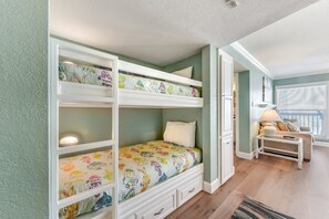 A bunk bed with floral bedding, a small bedside table with a lamp, and a view of a living area with a sofa and window blinds.