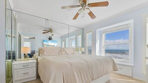 Une chambre à coucher comprenant un lit, une table de chevet, un ventilateur de plafond, une grande fenêtre donnant sur l’océan et un miroir.