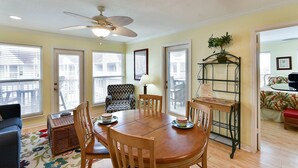 A dining area with a wooden table, chairs, and a ceiling fan. There are windows with blinds, a chair, and a small table with a lamp. - Seascape 2301 2 Bedroom Condo by RedAwning (Galveston)
