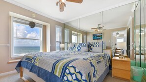 Ein Schlafzimmer mit einem großen Bett, einem Deckenventilator, einem Nachttisch, einem Spiegel und Blick aufs Meer.