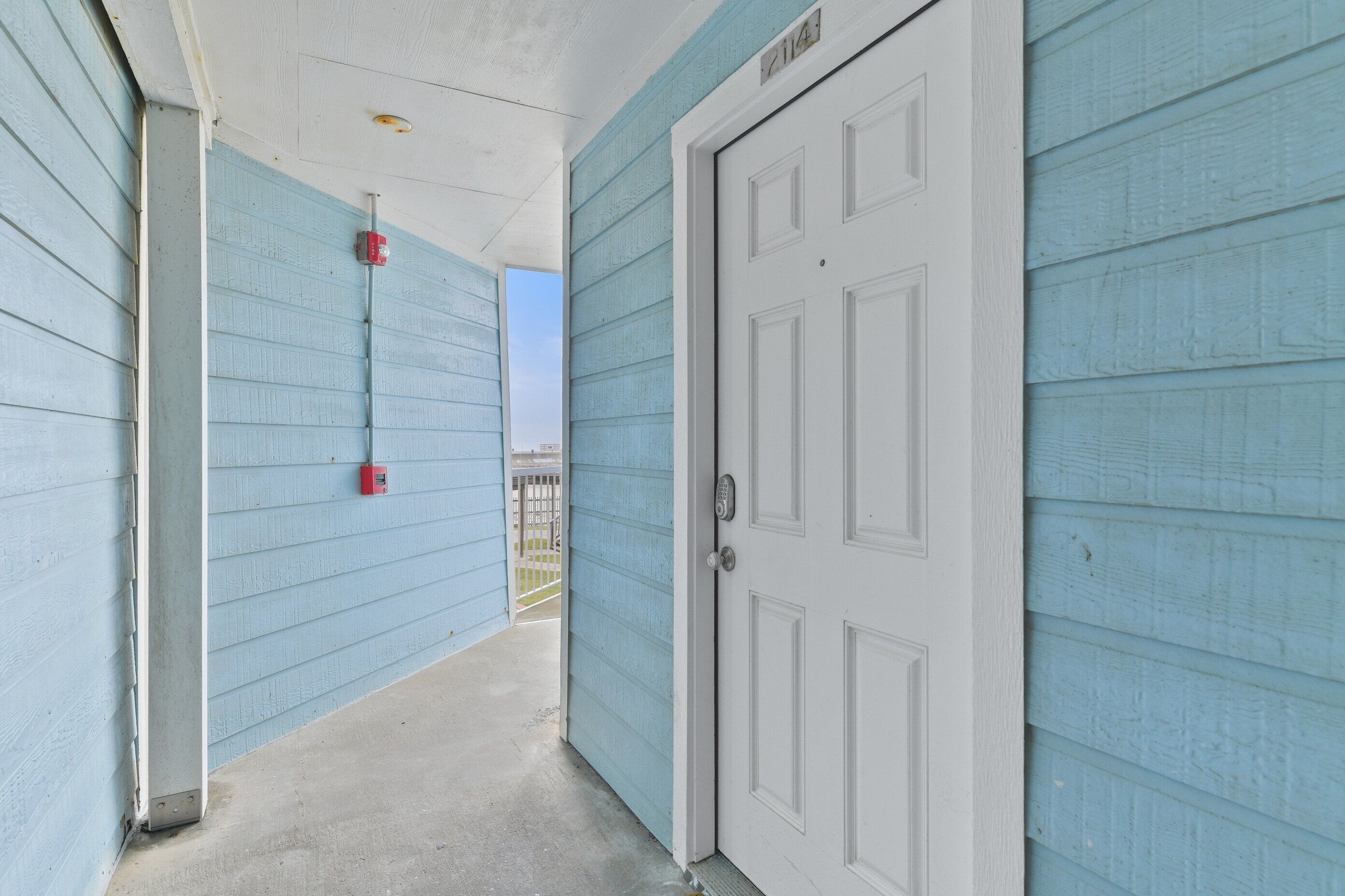 A narrow hallway with light blue walls, a white door with a number plate, and a red fire alarm box.