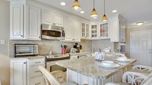 A kitchen with white cabinetry, a built-in microwave, a toaster, and a large island with seating. - Seascape 2113 2 Bedroom Condo by RedAwning (Galveston)