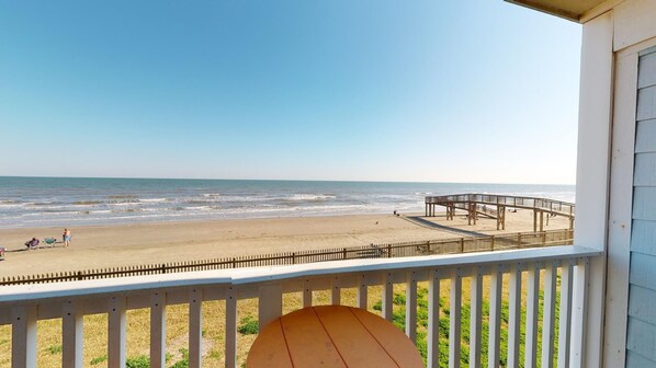 A beach view from a balcony with a wooden deck, a pier, and a sandy beach with people and beach chairs.