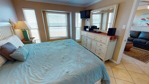 A bedroom with a bed, dresser, TV, and a view of a living area.