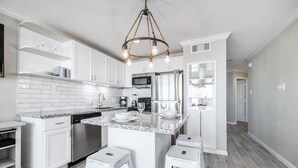 A modern kitchen with white cabinetry, a granite countertop, stainless steel appliances, and a central island with stools. - Seascape 1304 Studio Bedroom Condo by RedAwning (Galveston)
