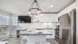 A modern kitchen with white cabinets, a granite countertop, stainless steel appliances, and a central island with stools. - Seascape 1304 Studio Bedroom Condo by RedAwning (Galveston)