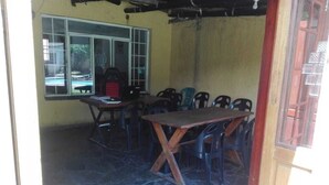 Une pièce avec un sol en pierre, une table en bois et des chaises en plastique. Il y a une fenêtre donnant sur une piscine.