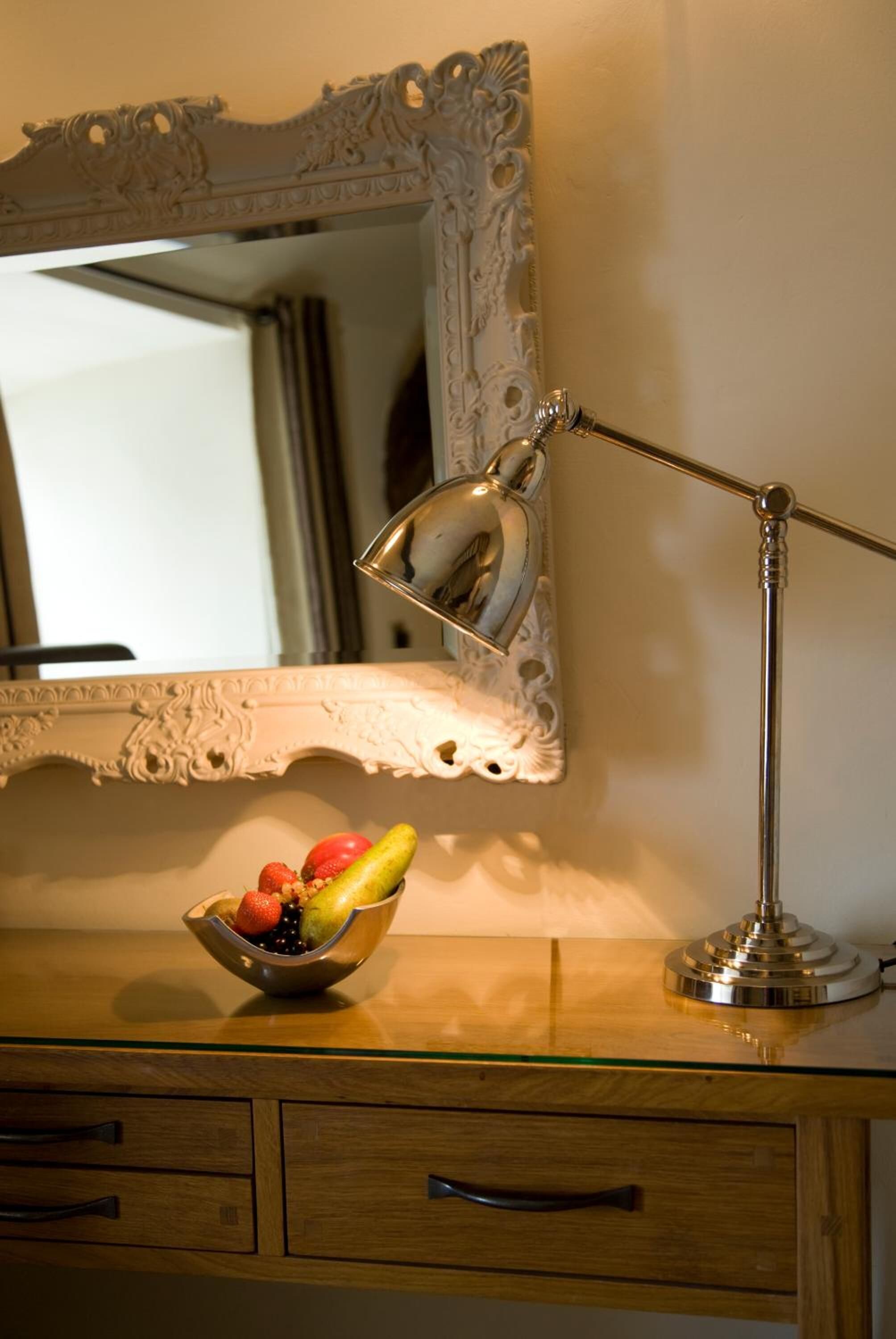 Un bureau en bois, un miroir, une lampe argentée et un saladier rempli de fruits.