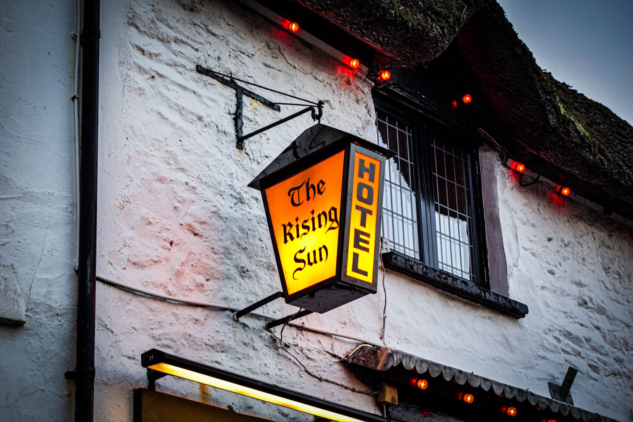 A hotel sign with the name "The Rising Sun Hotel" mounted on a white building with a window and a thatched roof.