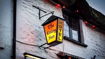 A hotel sign with the name "The Rising Sun Hotel" mounted on a white building with a window and a thatched roof.