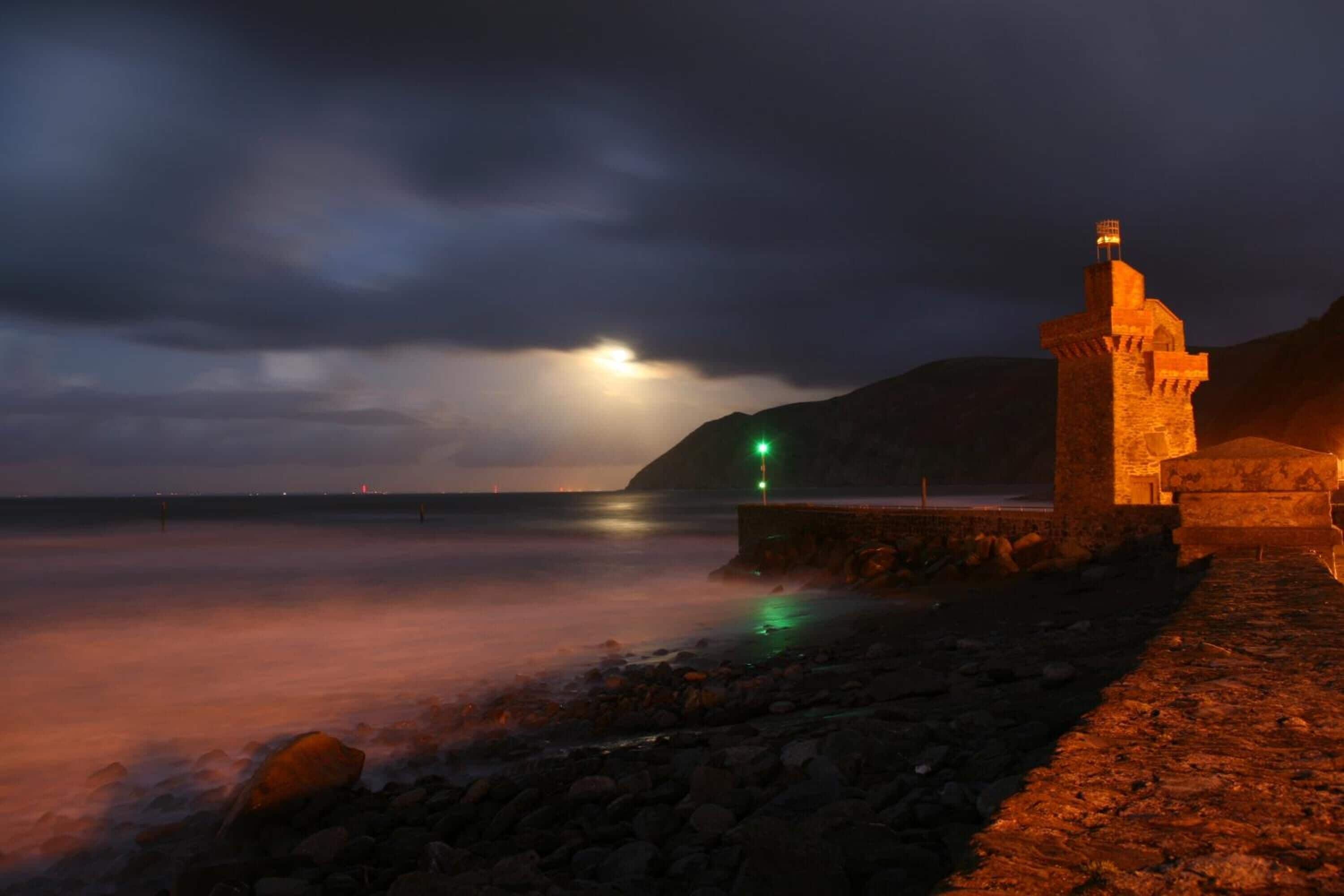 Uma torre de pedra à beira-mar à noite, sob um céu nublado.