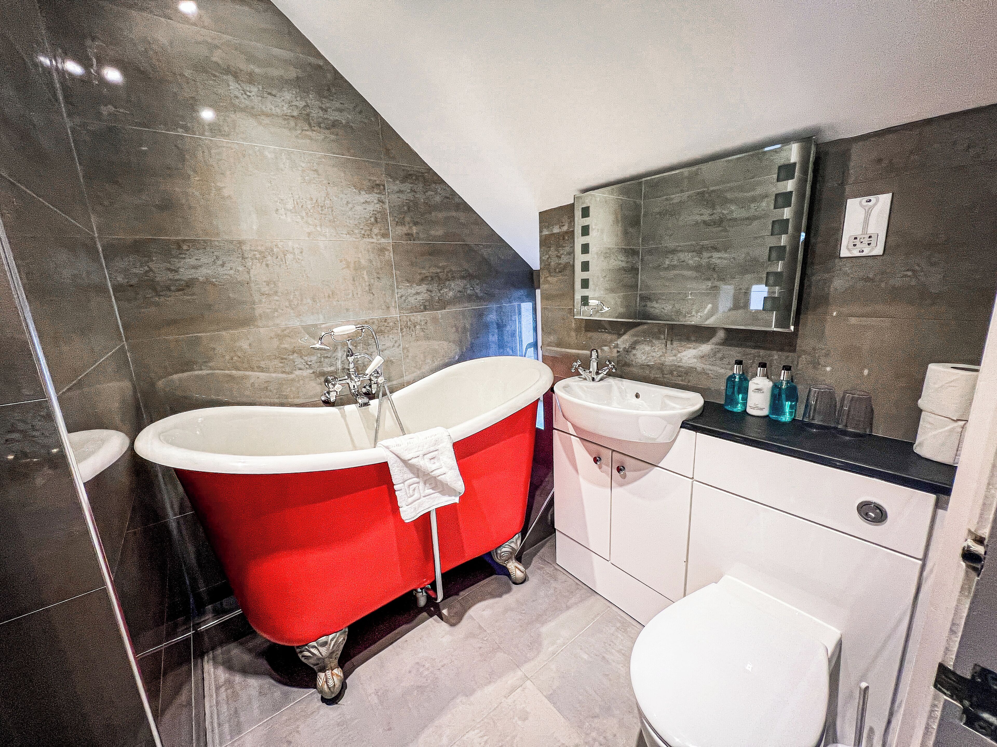 A modern bathroom with a red clawfoot bathtub, a white sink, and a toilet.