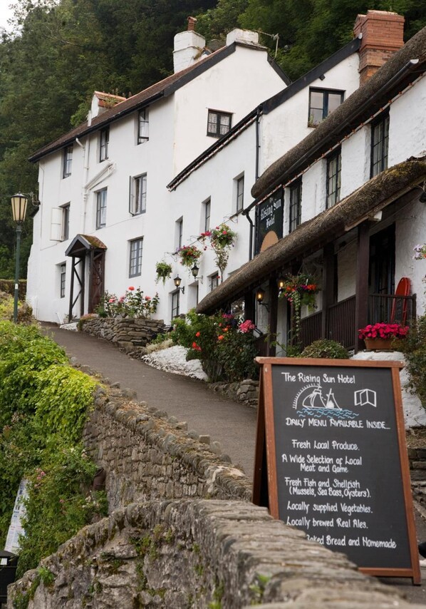 A white building with a signboard advertising The Rising Sun Hotel, offering fresh local produce, a variety of meats, seafood, locally supplied vegetables, and homemade bread.