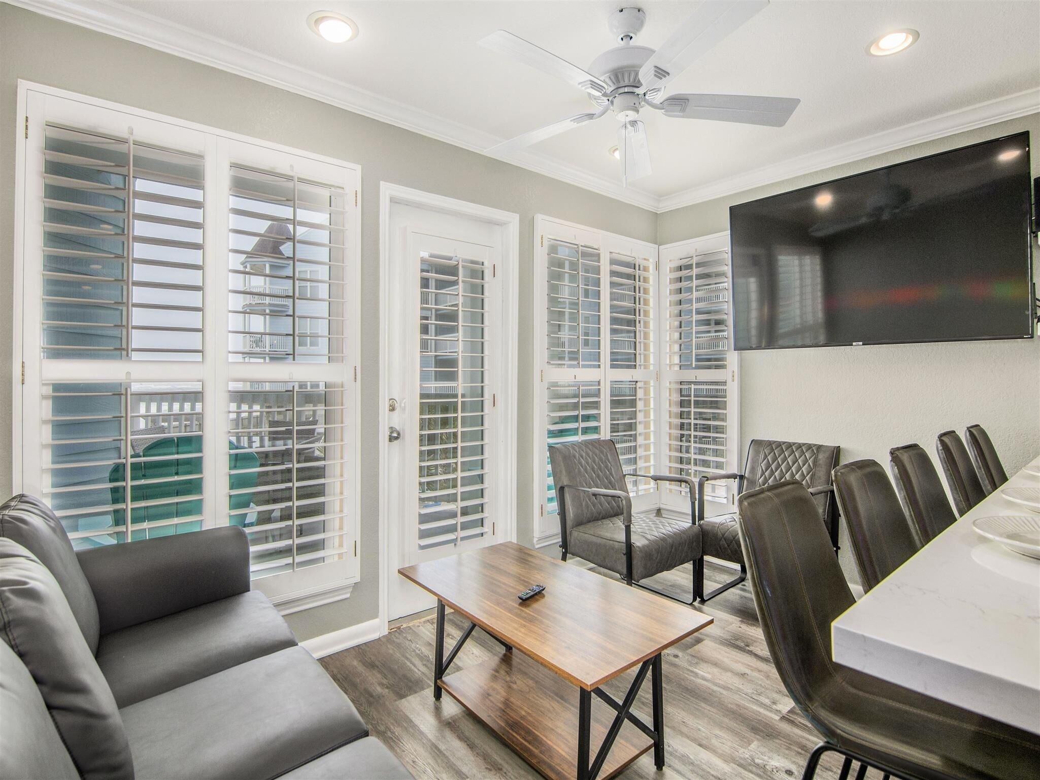 A modern living room with a dining area, large windows with blinds, a ceiling fan, and a flat-screen TV mounted on the wall.