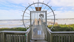 Une promenade en bois mène à un portail en métal orné d’un motif de palmier, d’où l’on a une vue sur une plage.