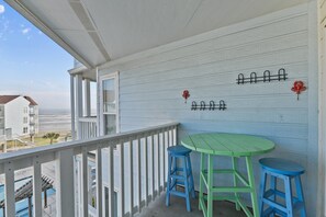Un balcon avec une table verte et des tabourets bleus, donnant sur une plage et des immeubles.