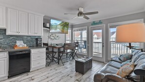 A kitchen with white cabinets, a dishwasher, and a dining area with a table and chairs. There is a ceiling fan, a TV mounted on the wall, and a sign that reads "Beach".