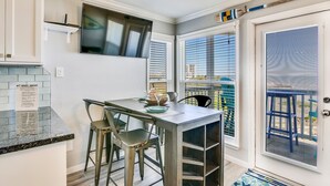 A kitchen with a dining area, a TV mounted above the counter, and a view of the outside through the window. - Seascape 2317 Studio Bedroom Condo by RedAwning (Galveston)